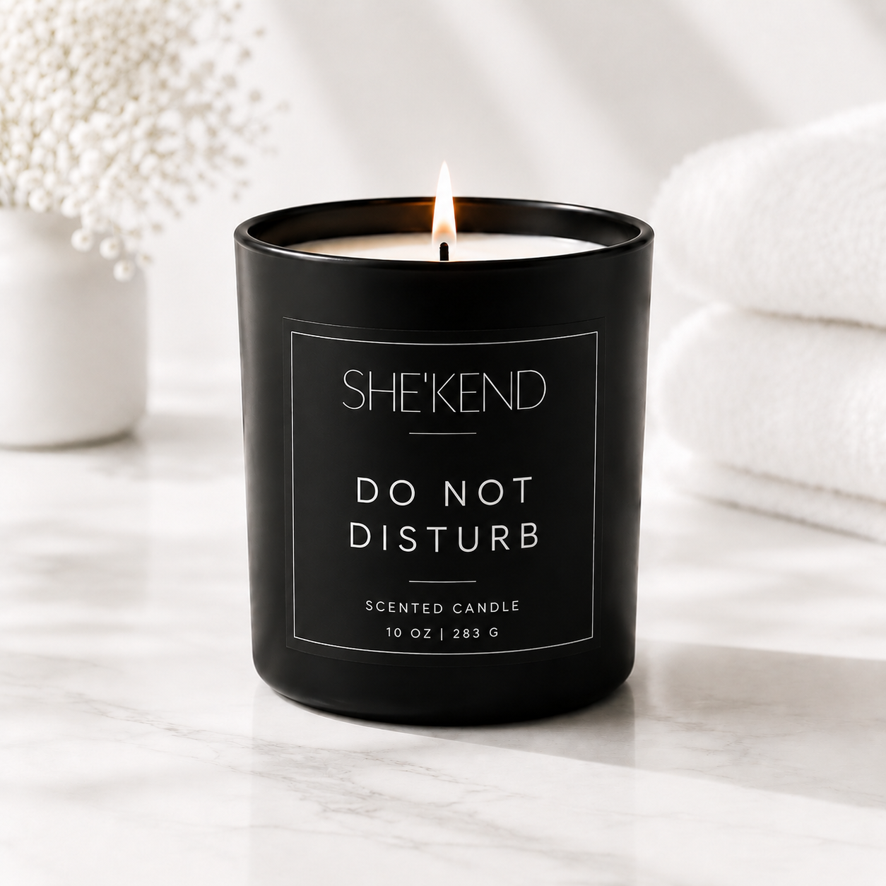 A black glass scented candle jar with a lit flame, labeled SHE'KEND DO NOT DISTURB, set on a white marble surface with folded white towels and dried flowers in the background.