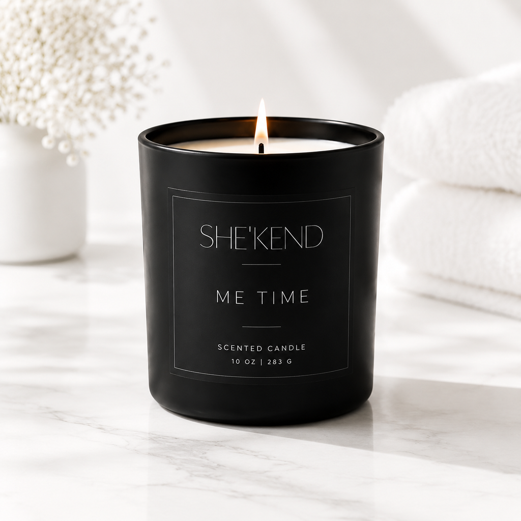 A black glass scented candle with a lit flame, labeled SHE'KEND ME TIME, set on a white marble surface with soft-focus white towels and dried flowers in the background.