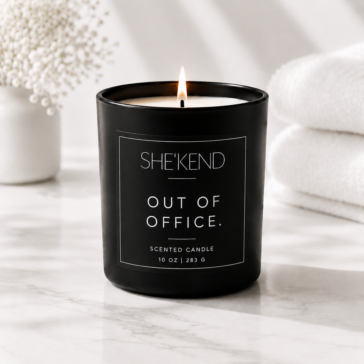 A black scented candle labeled SHE'KEND OUT OF OFFICE sits lit on a white marble surface, with soft-focus white towels and dried flowers in the background.