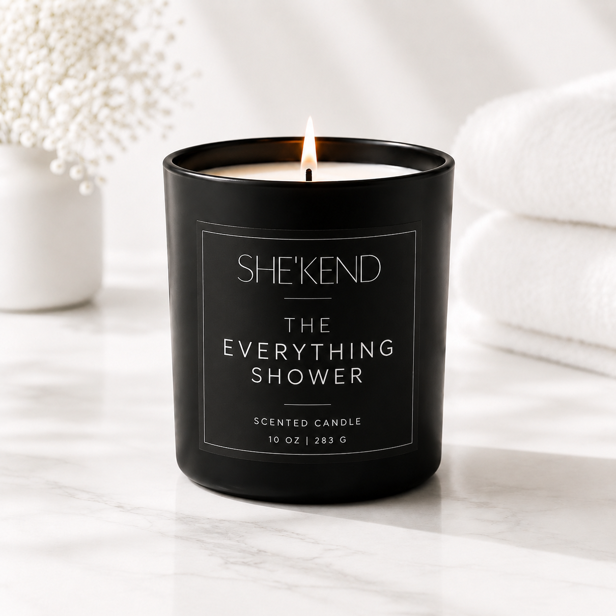 A black glass scented candle labeled SHE'KEND The Everything Shower, lit and set on a white marble surface with soft white towels and dried flowers in the background.